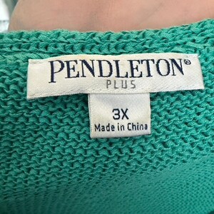 Puede incluir: Primer plano de una prenda de punto turquesa con una etiqueta que dice "PENDLETON PLUS" y "3X Made in China". La tela de punto tiene un aspecto texturizado. La etiqueta es blanca con texto negro.