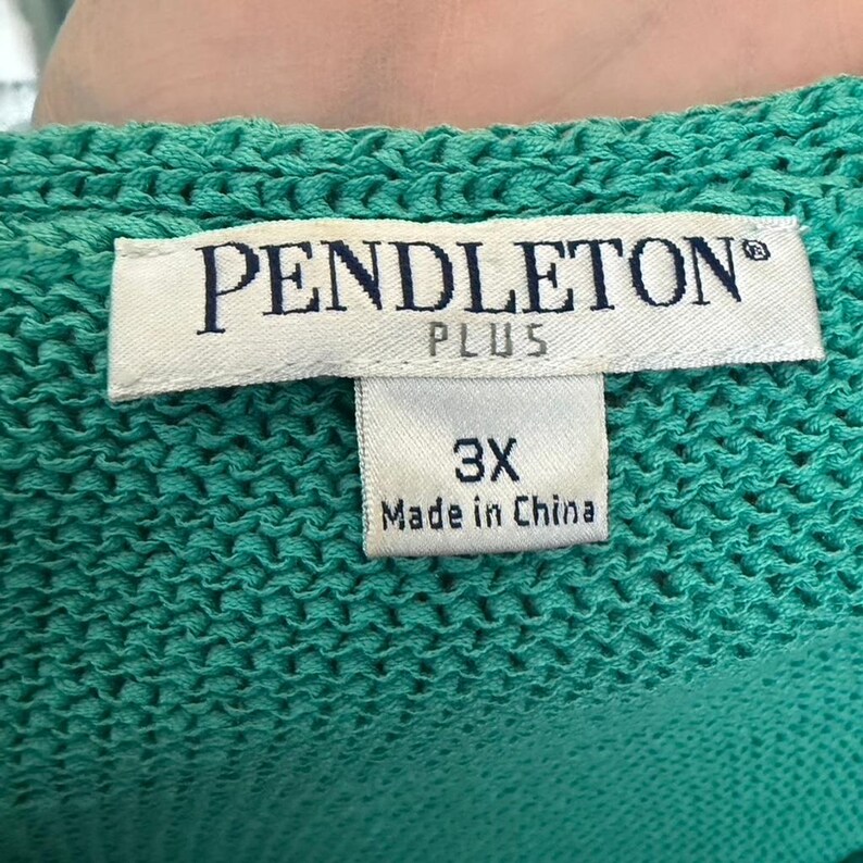Puede incluir: Primer plano de una prenda de punto turquesa con una etiqueta que dice "PENDLETON PLUS" y "3X Made in China". La tela de punto tiene un aspecto texturizado. La etiqueta es blanca con texto negro.