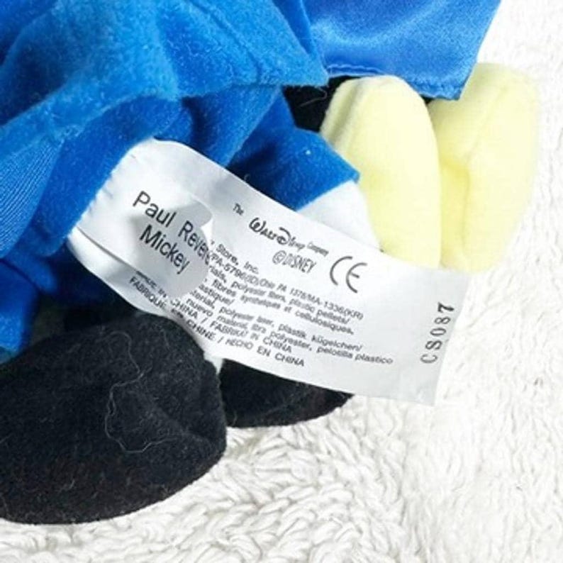 May include: A close-up of a Mickey Mouse plush toy. The toy is blue, black, and yellow. A white tag with the text "Paul Revere Mickey" is visible. The tag also includes the Disney logo and other text.