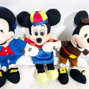 May include: Three Mickey Mouse plush toys in various outfits. The Mickey Mouse toys are wearing blue, red, and brown outfits with black ears and yellow shoes. The middle Mickey Mouse is wearing a colorful hat.