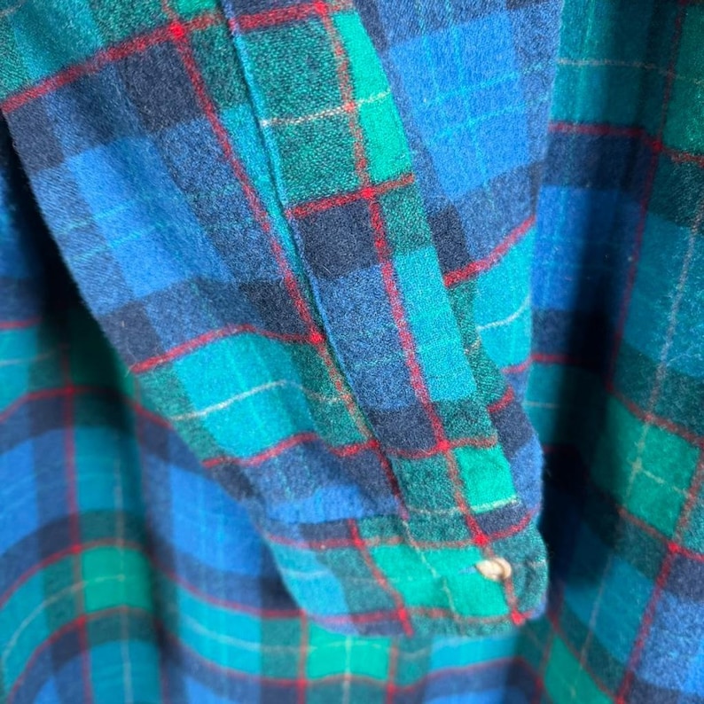 May include: Close-up of a blue, green, and red plaid flannel shirt. The fabric is a soft, woven material with a classic check pattern. The sleeve cuff is visible, with a button closure.