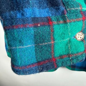 May include: Close-up of a section of a plaid garment. The fabric features a pattern of teal, green, and navy blue squares with thin red lines. A gold-colored button is visible on the edge of the fabric.