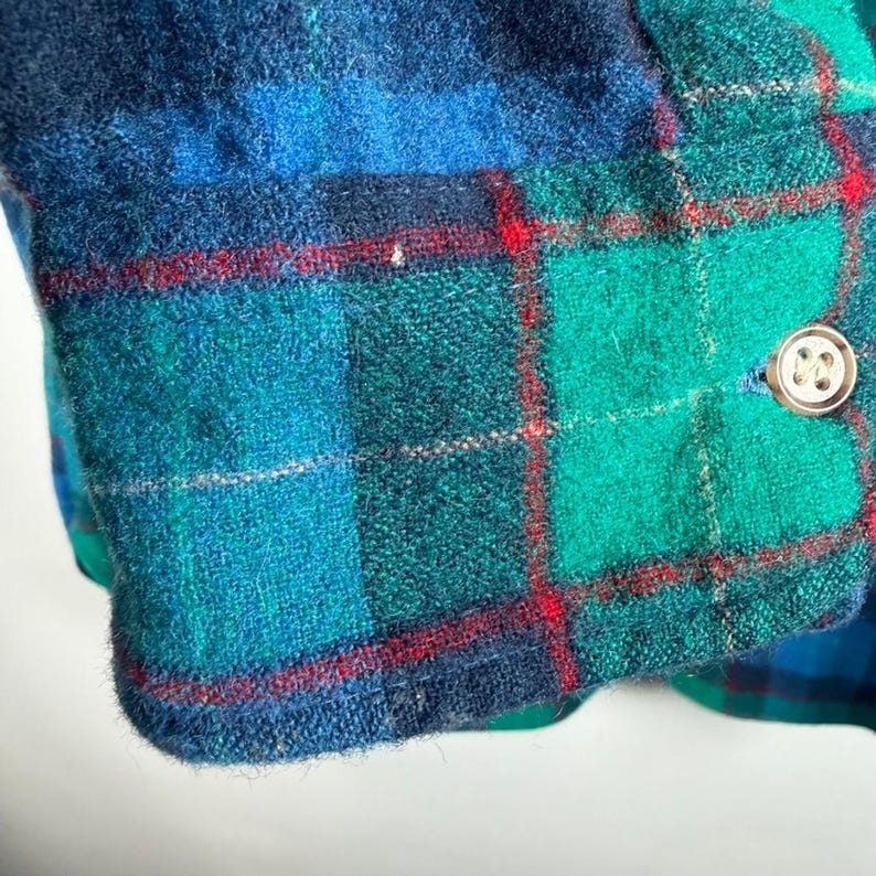 May include: Close-up of a section of a plaid garment. The fabric features a pattern of teal, green, and navy blue squares with thin red lines. A gold-colored button is visible on the edge of the fabric.