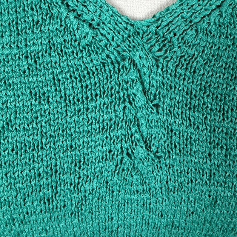 Puede incluir: Primer plano de un top de punto de color turquesa con cuello en V. La tela de punto tiene un aspecto texturizado con puntadas visibles y peque&ntilde;as aberturas. El escote presenta un detalle retorcido, a&ntilde;adiendo un elemento &uacute;nico a la prenda.