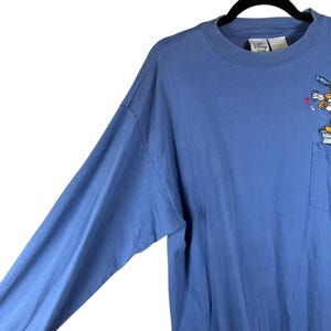 Pu&ograve; includere: Una t-shirt a maniche lunghe color blu pervinca con girocollo e taschino sul petto. La maglia presenta un'etichetta Disney Store all'interno del colletto e un piccolo ricamo raffigurante un personaggio dei cartoni animati sopra la tasca.