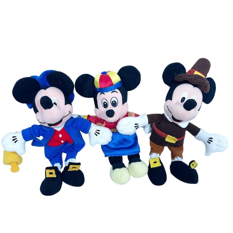 May include: Three Mickey Mouse plush toys in various outfits. One wears a blue suit with a red vest and holds a yellow bell. Another is dressed in a blue and red outfit with a yellow and red hat. The third wears a brown outfit with a pilgrim hat.