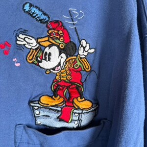 Pu&ograve; includere: Una maglia a maniche lunghe blu con un ricamo di Topolino vestito da direttore d'orchestra in un'uniforme rossa con dettagli dorati, in piedi su una piattaforma grigia mentre tiene una bacchetta, con piccole note musicali rosse e rosa.