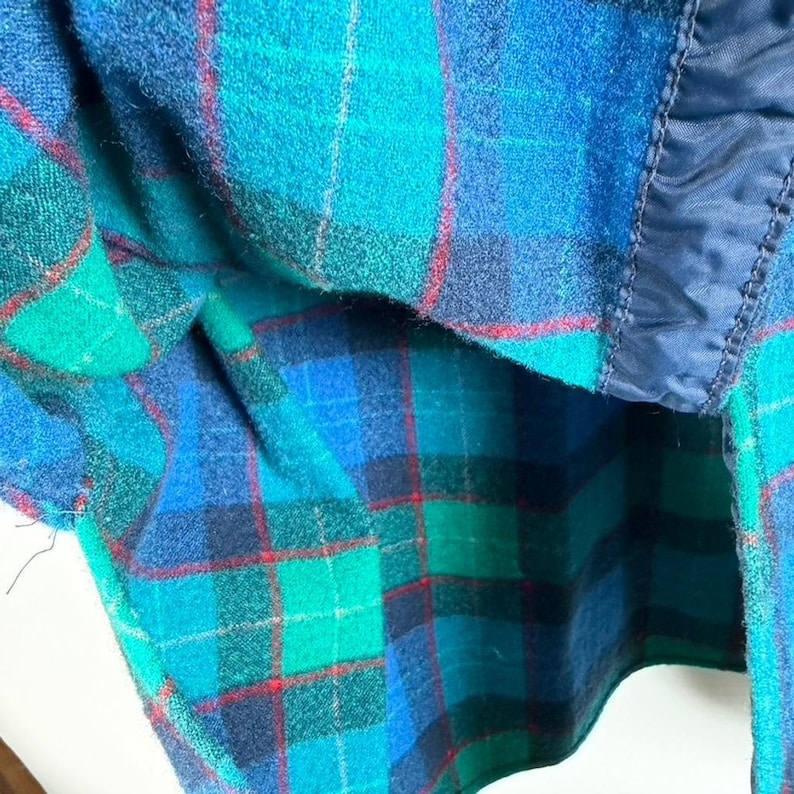 May include: Close-up of a plaid fabric in shades of blue, green, and red. The fabric has a textured appearance and a dark blue lining. The plaid pattern is visible throughout the image, with a dark blue trim.
