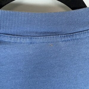 Pu&ograve; includere: Una vista ravvicinata di una t-shirt blu con scollo rotondo a coste. Il tessuto &egrave; di un colore blu solido di tonalit&agrave; media con una trama a maglia visibile. Il capo &egrave; esposto su una gruccia nera, mostrando chiaramente il colletto e il petto.