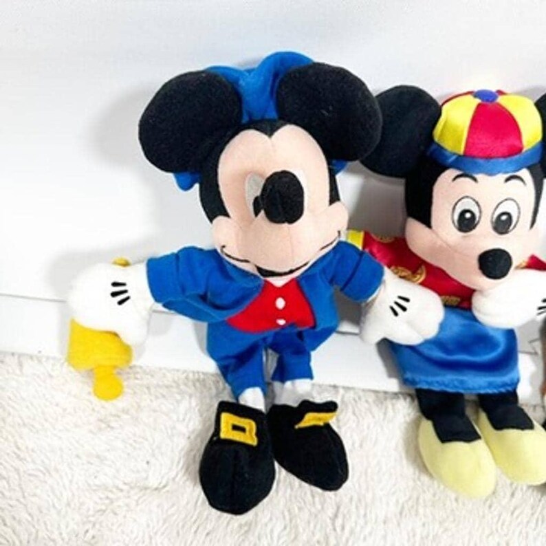 May include: Two Mickey Mouse plush toys. One Mickey is dressed in a blue jacket, red vest, and black shoes. The other Mickey wears a red and gold outfit with a blue skirt and a yellow, red, and blue hat.