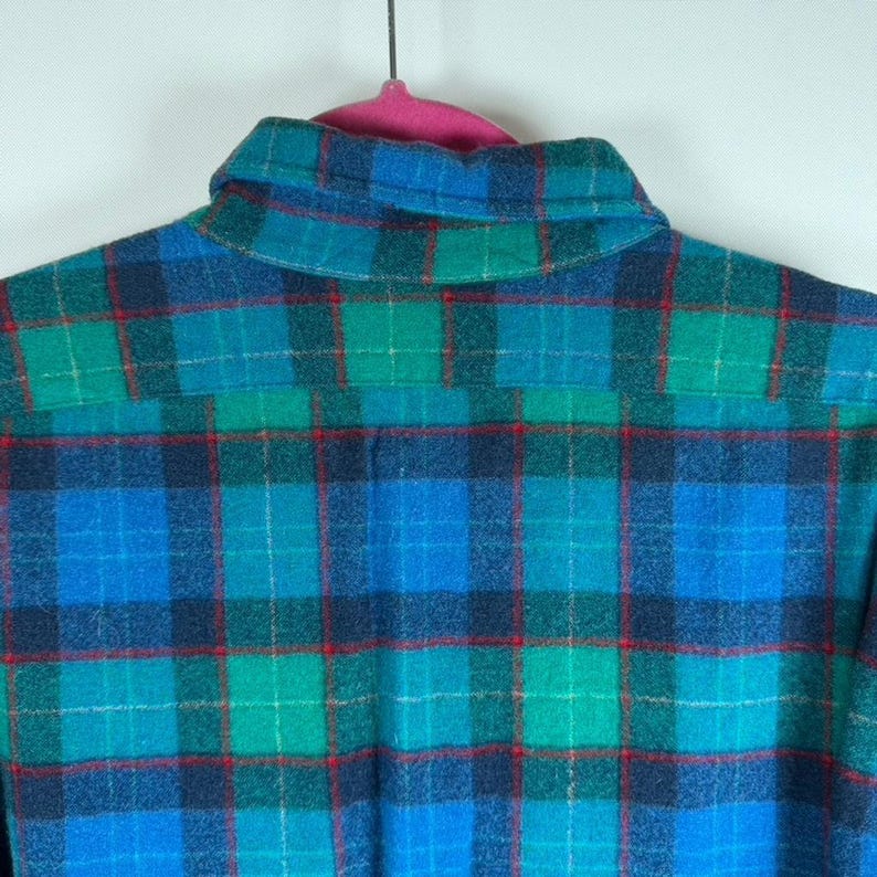 May include: A close-up of a blue, green, and red plaid flannel shirt. The shirt is hanging on a pink hanger. The plaid pattern is made up of intersecting lines of blue, green, and red, creating a classic look.