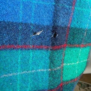 May include: Close-up of a blue, green, and navy plaid fabric with red accent lines. The fabric has a textured appearance and shows signs of wear with small holes. The plaid pattern is a classic design.