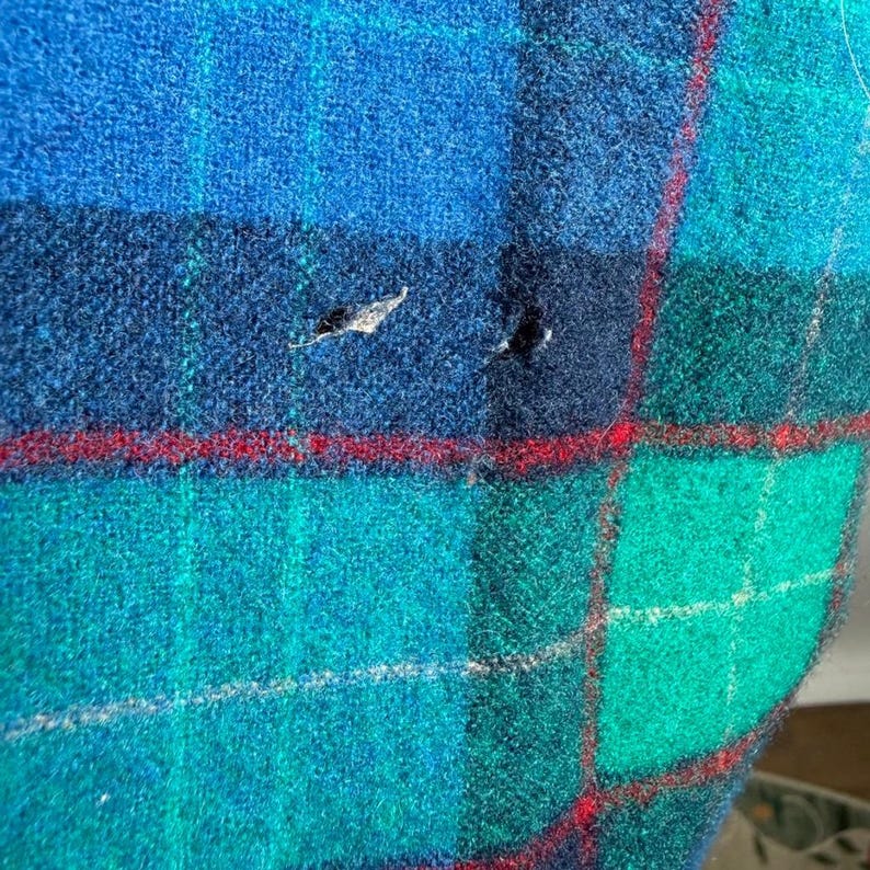 May include: Close-up of a blue, green, and navy plaid fabric with red accent lines. The fabric has a textured appearance and shows signs of wear with small holes. The plaid pattern is a classic design.