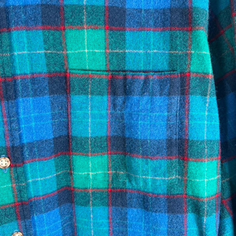 May include: Close-up of a blue, green, and red plaid flannel shirt. The shirt features a classic plaid pattern with a pocket and button details. The fabric appears soft and comfortable, suitable for casual wear.