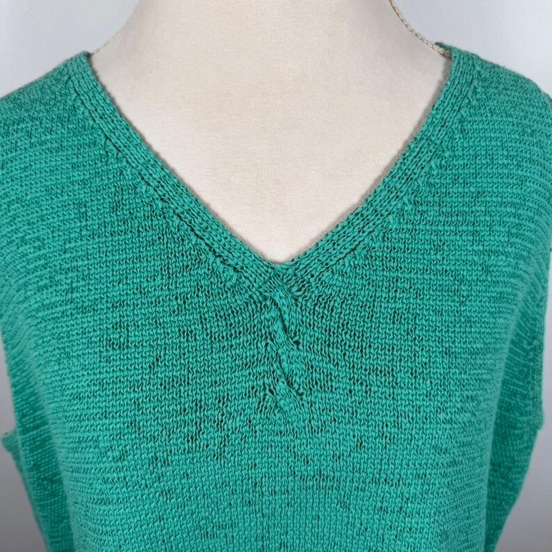 Puede incluir: Una camiseta sin mangas de punto verde esmeralda con cuello en V profundo. La tela de punto tiene una textura sutil y un ligero aspecto moteado. El escote est&aacute; reforzado con una costura en contraste, a&ntilde;adiendo un toque de detalle.