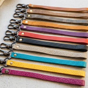 May include: A collection of leather wrist straps in various colors, including brown, gray, orange, purple, red, black, beige, teal, yellow, and magenta. Each strap has a metal clasp for attaching to a bag or other item.