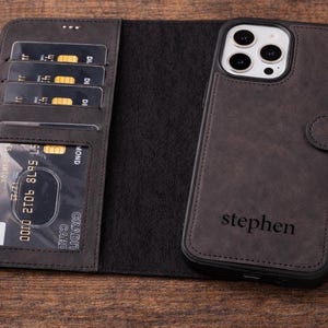 Może przedstawiać: Ciemnoszare skórzane etui na telefon z portfelem. Etui posiada magnetyczne zapięcie i miejsce na telefon. Sekcja portfela ma przegródki na karty kredytowe. Na etui wygrawerowano imię "stephen".
