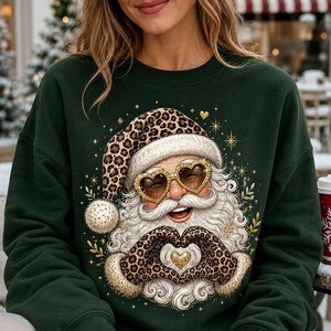 May include: A dark green crewneck sweatshirt featuring a graphic of Santa Claus wearing a leopard print hat and heart-shaped glasses. Santa has a white beard, is making a heart shape with leopard print gloved hands, and is surrounded by gold stars.