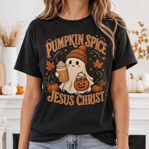 May include: A black short-sleeve t-shirt featuring a graphic of a ghost wearing a beanie, holding a pumpkin spice drink with a cross and a jack-o'-lantern bucket. The text PUMPKIN SPICE JESUS CHRIST is printed in orange above and below the ghost.