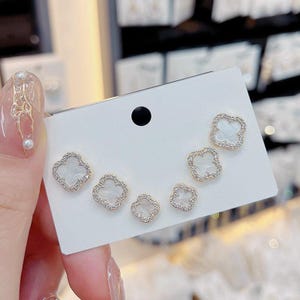 Può includere: Un set di sei orecchini a bottone a forma di quadrifoglio. Gli orecchini hanno un bordo color oro con accenti chiari e scintillanti. Il centro di ogni orecchino è bianco. Gli orecchini sono esposti su una carta bianca.