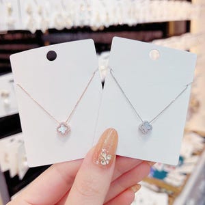 May include: Two necklaces with clover pendants, one in rose gold and the other in silver, presented on white cards. The pendants have a four-leaf clover design with a central white stone and clear crystal detailing. The necklaces are on a fine chain.