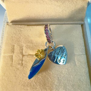 Op de afbeelding: Een zilveren bedel met een surfplank van blauw emaille en een goudkleurige bloem, gecombineerd met een hartvormige bedel gegraveerd met de tekst 'Hawaii' en 'Aloha'. Het oogje is bezet met kleine roze steentjes, een gedetailleerd sieraad.