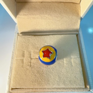 May include: A spherical charm featuring a vibrant yellow band, a bright red star at the center, and blue accents on the top and bottom. The charm is presented inside a cream-colored jewelry box with the brand name PANDORA printed in gold.