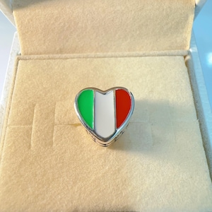 Könnte beinhalten: Ein herzförmiger Anhänger mit dem Design der italienischen Flagge, mit grüner, weißer und roter Emaille. Der Anhänger ist in einem silberfarbenen Metall gefasst und wird in einer beigen Schmuckschatulle präsentiert.