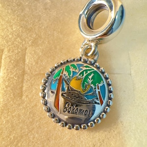 May include: A silver charm with a tropical scene featuring palm trees, a sunset, and the word "Bahamas." The charm has a beaded border and a loop for attaching to a bracelet or necklace. The design incorporates green, blue, orange, and yellow.
