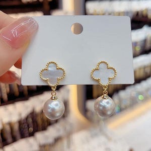 May include: Gold-coloured clover-shaped earrings with a white centre and a dangling pearl. The earrings are displayed on a white card, showcasing their elegant design and delicate details. The pearls are a soft, off-white colour.