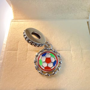Könnte beinhalten: Ein silberner Charm-Anhänger mit Fußball-Design. Der Ball hat rote, weiße, grüne und blaue Emaille in einem kreisförmigen Rahmen mit herzförmigen Details. Der Charm hat eine Schlaufe zum Befestigen.