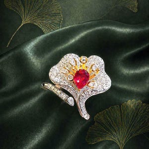 Puede incluir: Un anillo detallado con una piedra preciosa roja central en forma de corazón, rodeada de diamantes más pequeños y detalles dorados. El anillo está engastado en una banda plateada, y el diseño se asemeja a una flor o una hoja estilizada. El fondo es una tela verde oscuro y sedosa.