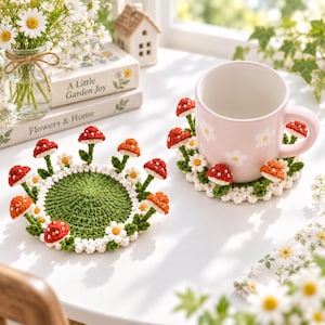 Può includere: Due sottobicchieri rotondi all'uncinetto con centro verde, bordo di margherite bianche e piccoli funghi tridimensionali rossi e arancioni. Uno è appoggiato in piano, l'altro sostiene una tazza in ceramica rosa chiaro con fiori bianchi.