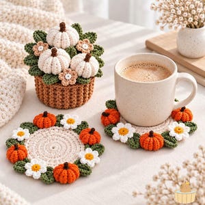Könnte beinhalten: Ein Set aus drei Häkelobjekten, bestehend aus einem braunen Korb mit weißen Kürbissen und kleinen Blumen sowie zwei runden Untersetzern, die mit orangefarbenen Kürbissen, weißen Gänseblümchen und grünen Blättern verziert sind.