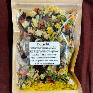 Peut inclure: Un sachet transparent rempli de légumes et d'épices séchés pour le bortsch. Le sachet est scellé et a un dos en papier brun. Une étiquette blanche fournit des instructions de préparation, y compris faire bouillir de l'eau et ajouter de la crème sure.