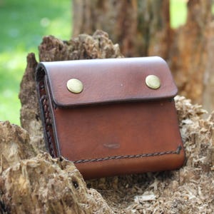 May include: A brown leather pouch with a rectangular flap secured by two brass-coloured metal snap buttons. The pouch features visible dark stitching along the bottom edge and sides, with a smooth, polished finish on the front surface.
