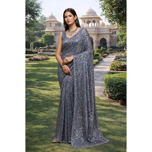 Könnte beinhalten: Ein grauer Pailletten-Sari mit einem ärmellosen Oberteil und drapiertem Stoff. Der Sari ist mit schimmernden Pailletten verziert, die einen glamourösen Look erzeugen. Die Frau trägt eine silberne Halskette und einen Ring. Der Hintergrund zeigt einen Garten und ein Gebäude.