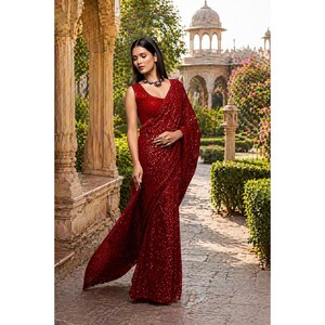 Könnte beinhalten: Eine Frau trägt einen roten Pailletten-Sari mit einem passenden ärmellosen Oberteil. Der Sari ist elegant drapiert und reflektiert das Licht. Der Hintergrund zeigt ein verziertes Gebäude und üppiges Grün.