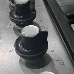 May include: Close-up of a stove top with black knobs. The knobs have a silver-colored top and are arranged in a row. The stove top is a light gray color.