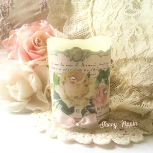 May include: A white candle with a floral design and a pink ribbon. The candle has a vintage look and is decorated with rhinestones. The text on the candle reads "Le mon bon amour a. Mamie" and "Rose Inturique".