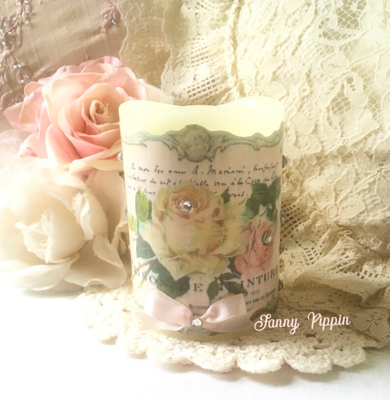 May include: A white candle with a floral design and a pink ribbon. The candle has a vintage look and is decorated with rhinestones. The text on the candle reads "Le mon bon amour a. Mamie" and "Rose Inturique".