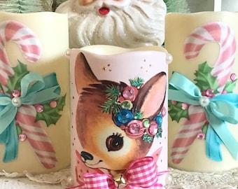 May include: Three white pillar candles decorated with a pink and white candy cane, a pink and white gingham bow with a bell, and a vintage image of a fawn wearing a pink gingham bow and a jeweled wreath.