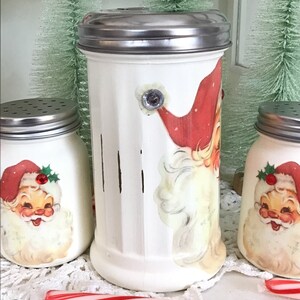 May include: Three white ceramic jars with Santa Claus designs and silver lids. The jars are decorated with vintage-style Santa Claus illustrations. The jars are sitting on a white doily with a red and white candy cane.