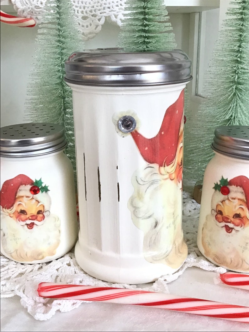 May include: Three white ceramic jars with Santa Claus designs and silver lids. The jars are decorated with vintage-style Santa Claus illustrations. The jars are sitting on a white doily with a red and white candy cane.