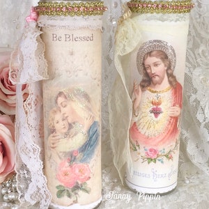 May include: Two decorative glass candles with religious imagery. One candle features an image of Mary and Jesus with the text "Be Blessed". The other candle depicts Jesus with a heart. Both candles are adorned with lace and pink accents.
