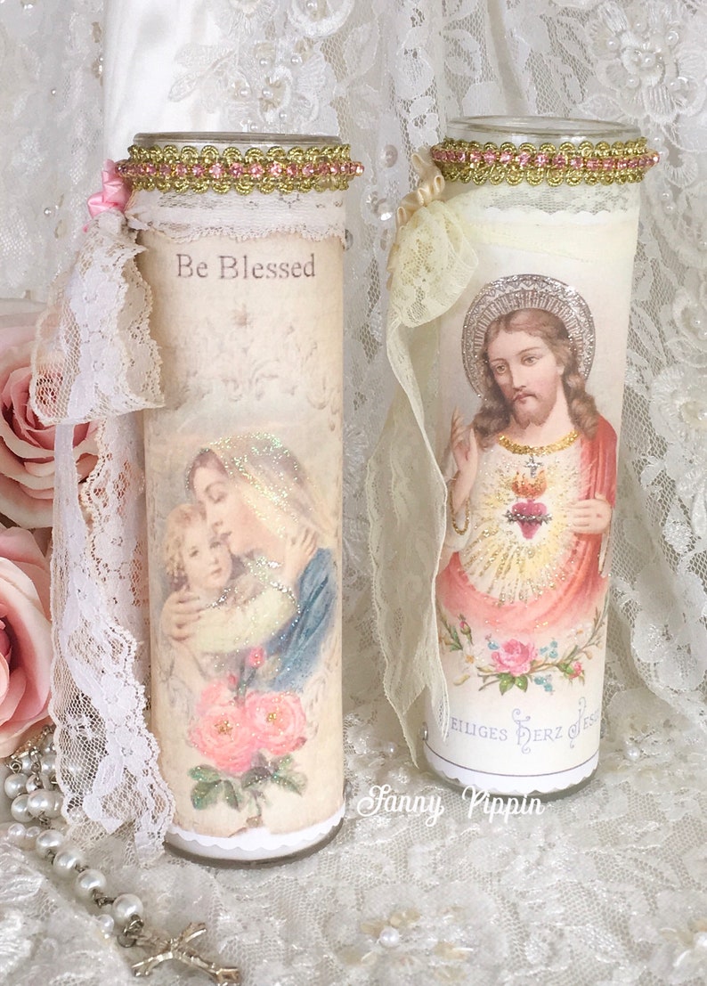 May include: Two decorative glass candles with religious imagery. One candle features an image of Mary and Jesus with the text "Be Blessed". The other candle depicts Jesus with a heart. Both candles are adorned with lace and pink accents.