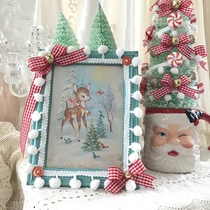 Puede incluir: Una escena navide&ntilde;a enmarcada con un ciervo, p&aacute;jaros y &aacute;rboles, acentuada con lazos de cuadros rojos y blancos, campanas y pompones. Un peque&ntilde;o &aacute;rbol de Navidad verde y una taza de Pap&aacute; Noel gui&ntilde;ando un ojo completan la exhibici&oacute;n festiva.