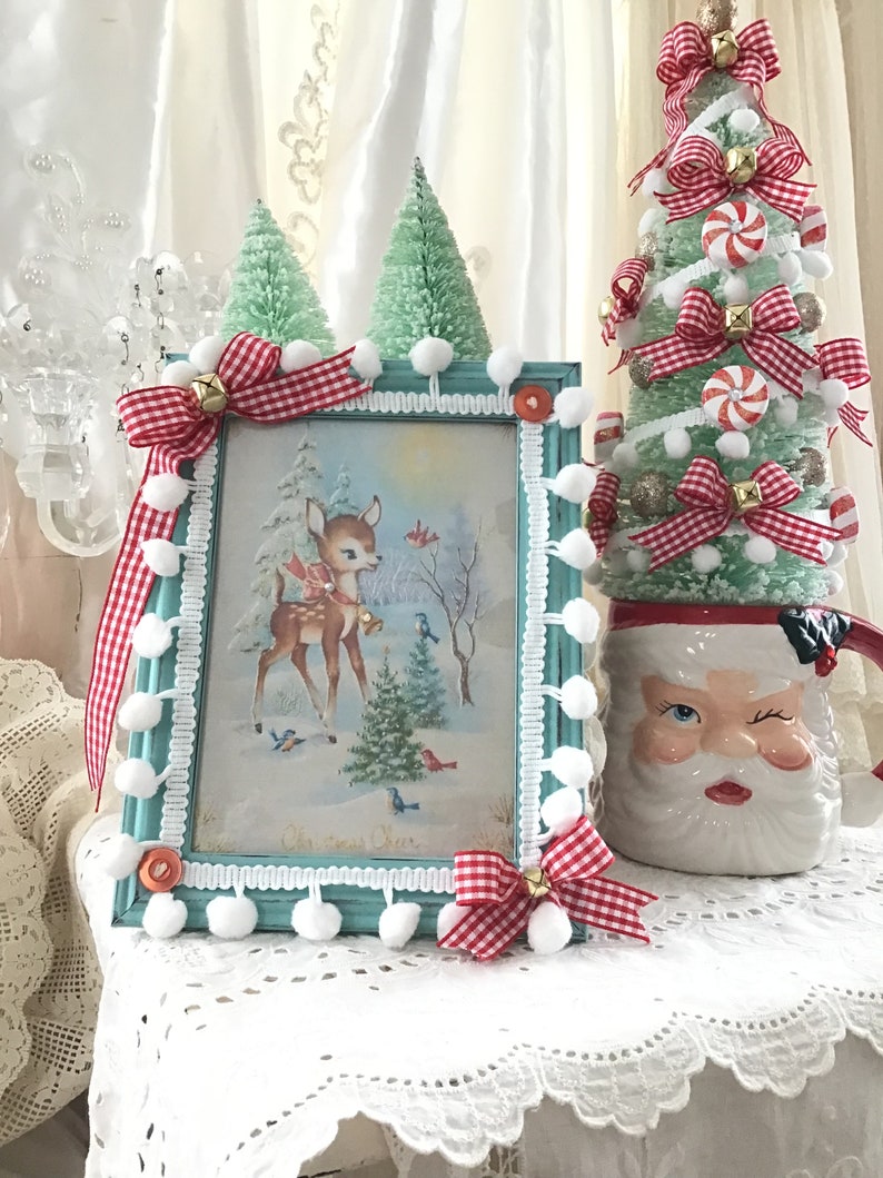 Puede incluir: Una escena navide&ntilde;a enmarcada con un ciervo, p&aacute;jaros y &aacute;rboles, acentuada con lazos de cuadros rojos y blancos, campanas y pompones. Un peque&ntilde;o &aacute;rbol de Navidad verde y una taza de Pap&aacute; Noel gui&ntilde;ando un ojo completan la exhibici&oacute;n festiva.