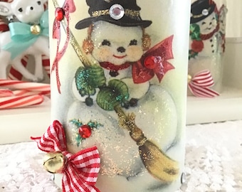 May include: A white ceramic candle holder with a vintage illustration of a snowman holding a broom. The snowman is wearing a black top hat with a gold band and a red scarf. The candle holder is decorated with red and white gingham ribbon and a gold bell.
