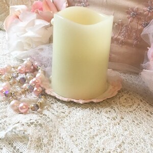 May include: A white pillar candle with a slightly curved top sits on a pink and white decorative plate. The candle is on a white lace tablecloth.